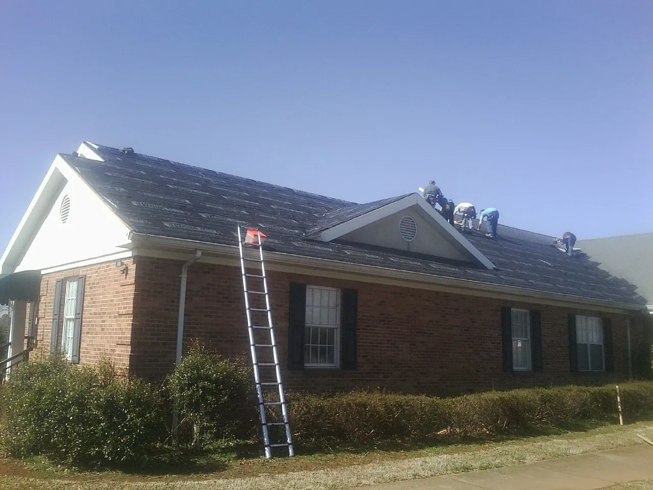 Crew tearing shingles off a brick ranch roof with a ladder set — Metal Roof Repair work by our crew in George